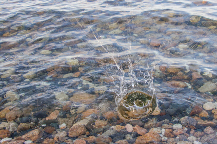Through clear flowing water, colored small stones are visible. There is pray of stone thrown into water.